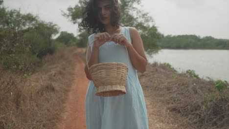 Slow-motion-handheld-shot-of-a-young-charming-indian-woman-in-a-light-blue-dress-with-a-wooden-basket-in-her-hands-standing-in-front-of-a-lake-in-nature-on-a-dried-footpath-looking-into-the-camera