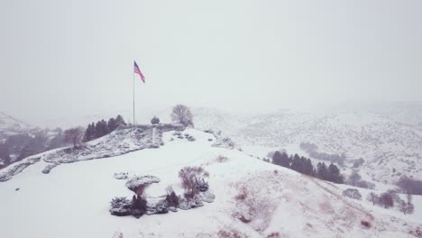 Aerial-shot-of-the-USA-flag-waving-on-top-of-a-snow-covered-landscape-during-winter
