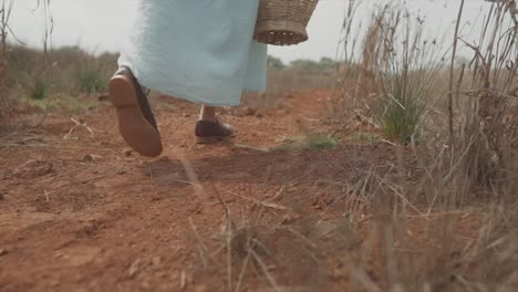 Close-up-of-a-woman-in-a-blue-dress-with-a-basket-in-her-hand-walking-across-a-dirt-field