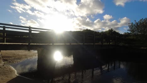 Sunset-time-lapse-clouds-mirrored-under-wooden-bridge-flowing-beach-creek
