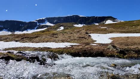 Snow-melting-rapidly-during-hot-spring-day-and-a-fresh-cold-river-is-passing-close-in-front-of-camera---Remaning-snow-cape-on-top-of-Finnbu-mountain-at-vikafjell-area-in-background---Static-Norway