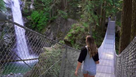 Woman-crossing-old-traditional-bridge-in-forest-in-British-Columbia-BC,-Canada-with-waterfall-and-river