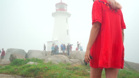 Young-girl-walking-towards-white-lighthouse-in-the-fog-on-an-overcast-day-in-Nova-Scotia,-Canada-in-red-dress
