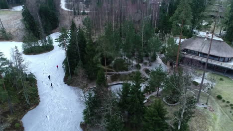 Aerial:-friends-having-fun-skiing-on-a-frozen-lake-in-front-of-a-cottage-into-a-forest-with-tall-trees