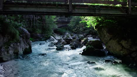 Aerial-Flying-along-forest-mountain-stream-rapids-under-wooden-small-bridge