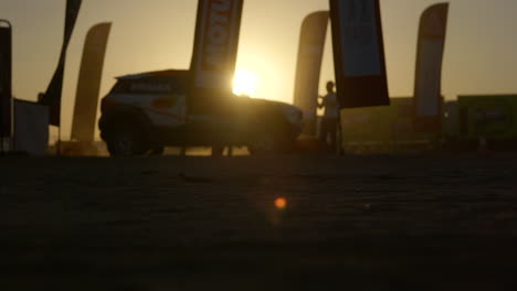 Silhouette-Von-Rennwagen-Und-Lkw,-Die-In-Zeitlupe-Auf-Einem-Sonnenuntergang-Im-Dakar-rallye-camp-Fahren