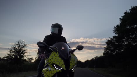Biker-man-on-BMW-sport-motorcycle,-young-man-rider-on-trendy-motorbike-stopped-on-asphalt-road-in-countryside-to-rest-during-the-trip-at-sunset---orbiting-gimbal-shot