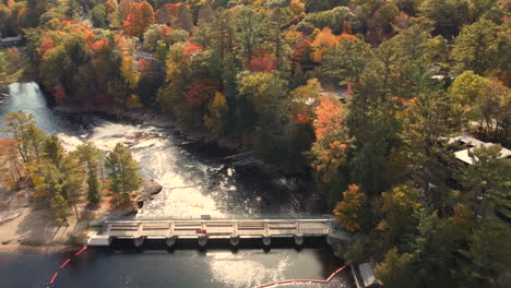Beautiful-landscape-view,-aerial-flyover-Port-Sydney-Falls,-capturing-golden-sunlight-reflection-on-the-water-surrounded-by-vibrant-and-colorful-tree-foliages-in-autumn-at-Huntsville,-Ontario-Canada