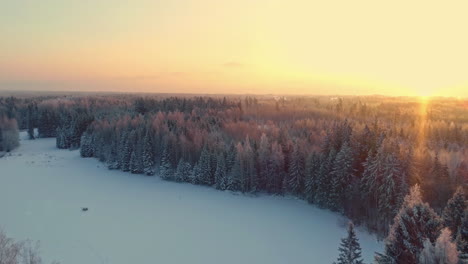 Aerial-drone-backward-moving-shot-over-frozen-lake-surrounded-by-coniferous-forest-covered-with-white-snow-at-sunrise