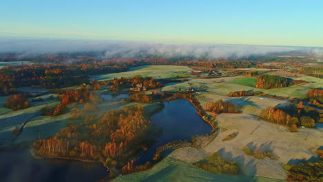 Aerial-flyover-natural-lake-surrounded-by-autumnal-forest-landscape-with-fog-and-frozen-grass