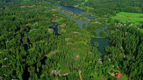 Verdant-landscape-with-small-isolated-houses-immersed-in-lush-nature-of-forest-and-lakes,-aerial-flyback-drone-view
