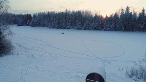 Aerial-drone-backward-moving-shot-over-wooden-cottage-and-barrel-sauna-on-a-white-snow-covered-landscape-during-morning-time