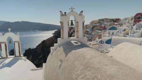 Panorámica-Sobre-Coloridas-Flores-Y-Campanas-De-La-Iglesia-Griega-Con-Fondo-De-Vistas-Al-Mar-En-Santorini,-Grecia