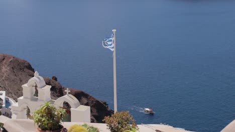 Campanas-De-La-Iglesia-Griega-Y-Bandera-Con-Mar-Azul-Y-Barco-En-El-Fondo-En-Santorini,-Grecia