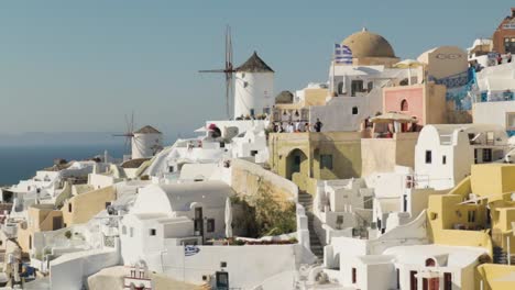 Close-pan-reveal-of-white-traditional-windmills-and-greek-flag-in-Santorini,-Greece
