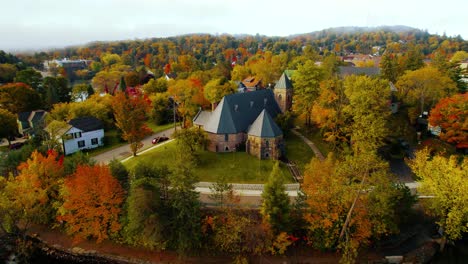 Drone-aerial-view-of-historic-cathedral-church-surrounded-by-village-homes-and-vibrant-Autumn-trees,-Huntsville-Ontario