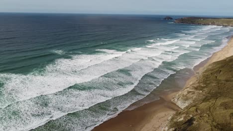 A-drone-shot-of-a-stunning-Cornish-beach,-revealing-a-vast-length-of-golden-sand-with-surfing-waves-coming-ashore-and-a-beautiful-headland-in-the-distance
