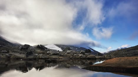 Timelapse-of-clouds-partly-covering-a-mountain-in-Norway,-with-pond-in-foreground