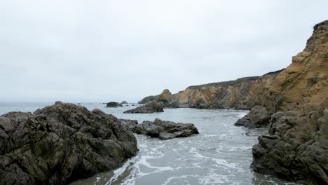 Flying-low-over-the-ocean-in-San-Mateo-County-beach-over-rocks-and-waves,-California