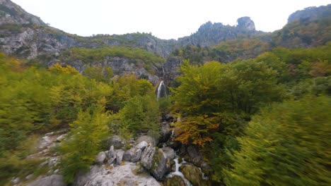 Flight-up-a-mountain-river-to-a-waterfall-through-autumn-coloured-forest-in-Theth,-Albania