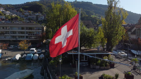 Fantástica-Toma-Aérea-En-Acercamiento-A-La-Bandera-De-Suiza,-Toma-Realizada-En-El-Municipio-De-Oberhofen-En-Suiza,-En-Un-Día-Soleado