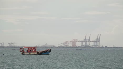 A-moored-Thai-fishing-boat-in-the-middle-a-bay,-in-the-background-the-cranes-of-the-Sri-Rascha-deep-sea-port,-Thailand