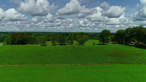 Aerial-drone-backward-moving-shot-over-green-farmland-along-with-hay-bales-scattered-out-on-the-field-with-white-clouds-passing