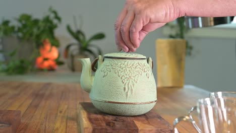 Making-herbal-tea-in-a-green-teapot-on-a-wood-table-in-a-light-and-airy-room-with-green-plants-in-the-background-and-a-glass-teacup