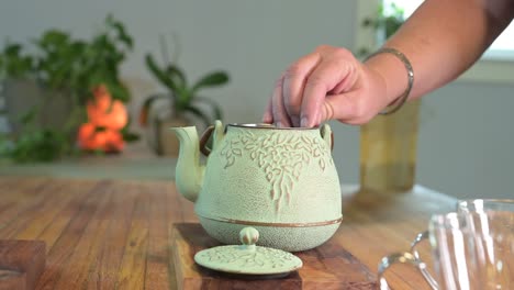 Making-herbal-tea-in-a-green-teapot-on-a-wood-table-in-a-light-and-airy-room-with-green-plants-in-the-background-and-a-glass-teacup