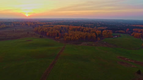 Sunset-aerial-pullback-over-vast-green-meadows-next-to-forested-landscape-with-canopy-in-fall-colors---beautiful-golden-hour-scene-with-orange-horizon