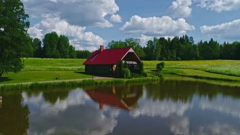 Wooden-hut-with-red-roof-on-lake-banks-in-bucolic-and-idyllic-green-landscape