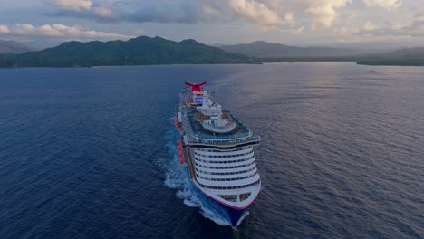 Aerial-orbit-shot-of-cruising-cruise-ship-with-luxury-pools-and-tv-monitor-on-rooftop-in-Dominican-Republic---Sunset-time-and-clouds-at-sky