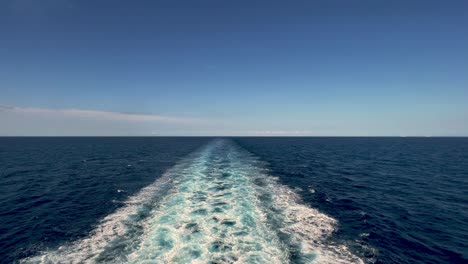Stern-view-of-wide-water-wake-left-from-cruise-ship-on-sea-water-surface-with-horizon-in-background-and-shadow-of-people-leaning-over-parapet-of-deck