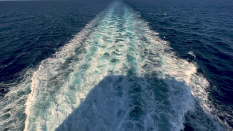 Rear-view-of-wide-water-wake-left-from-cruise-ship-on-sea-water-surface-with-horizon-in-background-and-shadow-of-people-leaning-over-parapet-of-deck