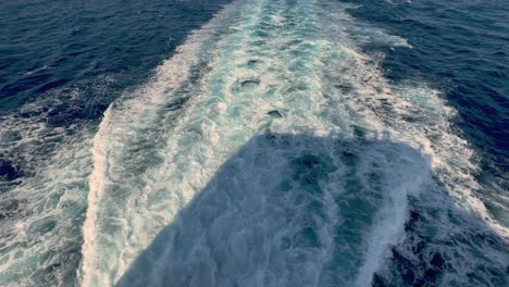 Back-view-of-wide-water-wake-left-from-cruise-ship-on-sea-water-surface-with-horizon-in-background-and-shadow-of-people-leaning-over-parapet-of-deck