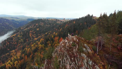 Aerial-orbit-of-a-couple-stand-on-top-of-a-mountain-valley-peak,-fall-color