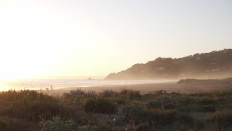 Zeitlupenschwenk-Eines-Wunderschönen-Strandes-Mit-Pflanzen,-Sand,-Den-Hügeln-Im-Hintergrund-Und-Dem-Meer,-Das-Den-Sonnenuntergang-Auf-Dem-Wasser-Widerspiegelt