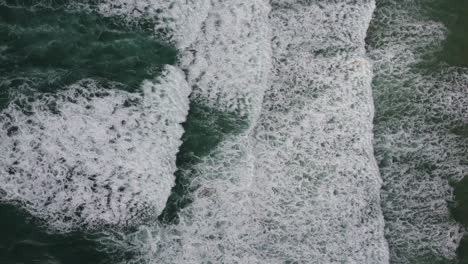 A-top-down-cinematic-drone-shot-of-green-Cornish-waves,-ideal-for-surfing,-slowly-rolling-and-breaking-on-the-shore