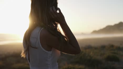 Static-slow-motion-handheld-shot-of-a-woman-in-a-white-dress-brushing-through-her-hair-watching-the-beautiful-sunset-on-the-beach