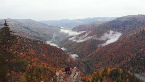 Aerial-view-couple-stand-on-top-of-a-rock-near-mountain-valley,-autumn