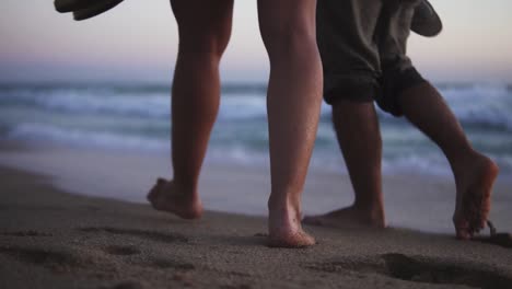 Slow-motion-low-angle-handheld-shot-of-a-couple-in-love-walking-at-dusk-on-a-beautiful-sandy-beach-in-front-of-the-sea-with-calm-waves