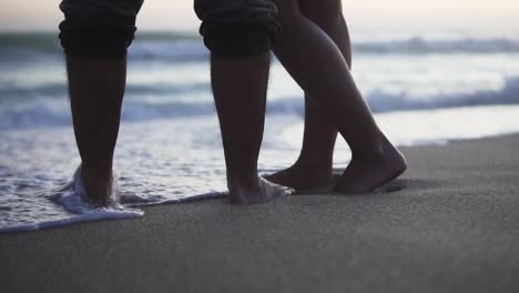 Static-slow-motion-shot-of-a-couple-in-love-standing-in-the-sand-on-the-beach-with-calm-waves-from-the-sea-during-a-romantic-sunset