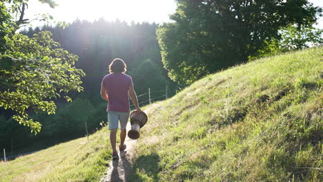 Young-man-with-red-Tshirt,-long-brown-hair-and-drum-in-his-hand-is-walking-on-a-small-path-on-a-beautiful-meadow-towards-the-sunset
