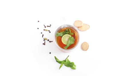 lemon-tea-in-a-transparent-glass-cup-with-sliced-lemon-and-ginger-mint-leaves-cloves-cardamom-black-pepper-in-white-background-rotating-top-shot
