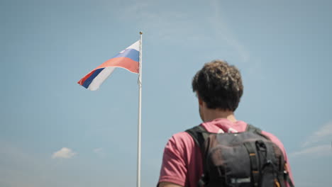 Ein-Junger-Wanderer,-Der-Seinen-Blick-Vor-Der-Sonne-Schützt-Und-Auf-Den-Mast-Mit-Der-Slowenischen-Flagge-Blickt,-Die-Im-Wind-Flattert