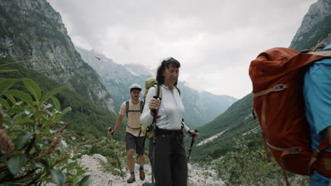 Hikers-passing-the-camera-to-the-right,-looking-into-the-green-valley-and-high-mountains-with-couds-surrounding-the-peaks