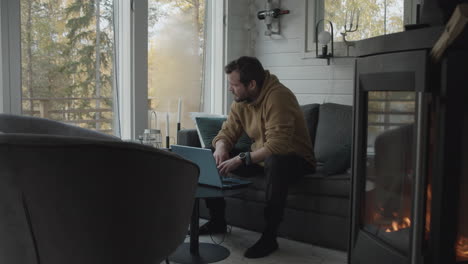 Man-with-laptop-works-daytime-job-in-front-of-fireplace-in-cozy-small-cabin-home