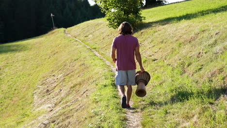 Young-man-with-red-Tshirt,-long-brown-hair-and-drum-in-his-hand-is-walking-on-a-small-path-on-a-beautiful-meadow