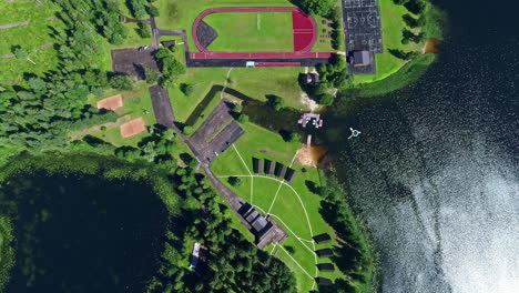 Top-Down-Ansicht-Sportkomplex-In-üppigen-Rasenflächen-Am-Wasser,-Absteigende-Drohnenaufnahme