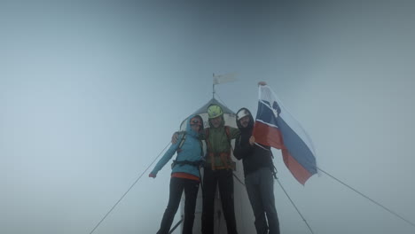 Hikers-on-the-top-of-a-mountain-Triglav-next-to-the-Aljaž-Tower-with-a-Slovenian-flag-happy-looking-at-the-camera